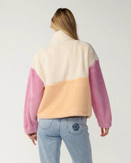 A woman viewed from behind wearing a fuzzy color-block fleece jacket with cream, peach, and pink sections, paired with light blue jeans against a plain light gray studio background.