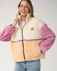 A woman wearing a sherpa fleece zip-up jacket with a cream upper section, pink sleeves, and a peach lower section, paired with light blue jeans against a light gray studio background.