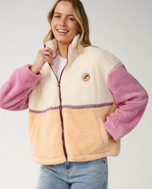 A woman wearing a sherpa fleece zip-up jacket with a cream upper section, pink sleeves, and a peach lower section, paired with light blue jeans against a light gray studio background.
