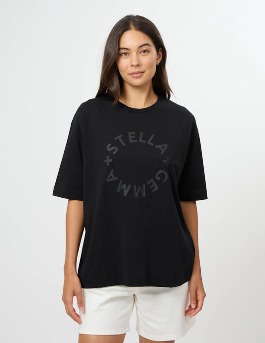 A woman modeling a black short-sleeve t-shirt with a subtle dark gray circular 'STELLA GEMMA' graphic on the front, paired with white shorts, against a plain light gray background.