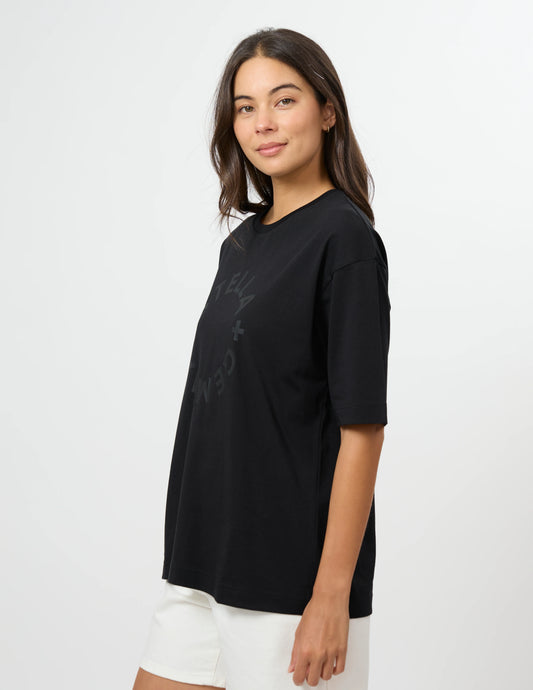 A woman wearing a black oversized short-sleeve t-shirt with a subtle tonal graphic on the front, paired with white shorts, photographed against a clean white studio background.