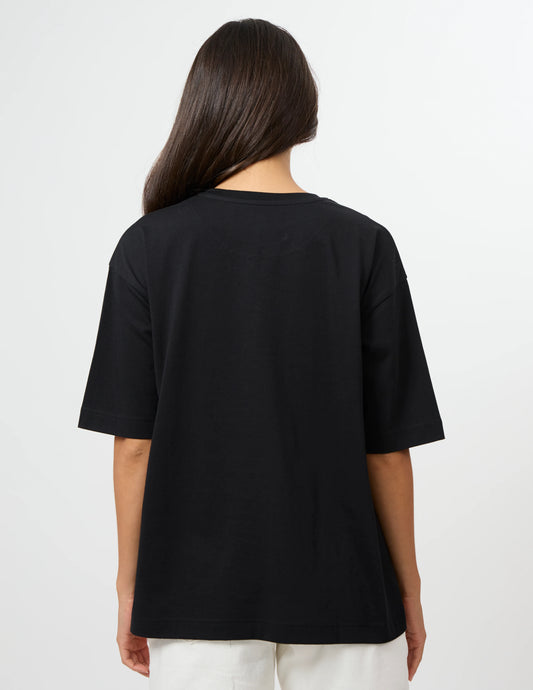 A woman viewed from the back wearing an oversized black short-sleeve T-shirt and white pants against a plain light gray studio background.