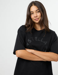 A woman with long dark brown hair wearing a black short-sleeve t-shirt with a subtle dark gray 'STELLA' graphic across the chest, posed against a light gray studio background.