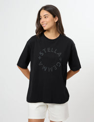 A woman modeling an oversized black t-shirt with a dark gray circular 'STELLA GEMMA' graphic on the chest, paired with white shorts, against a plain white studio background.