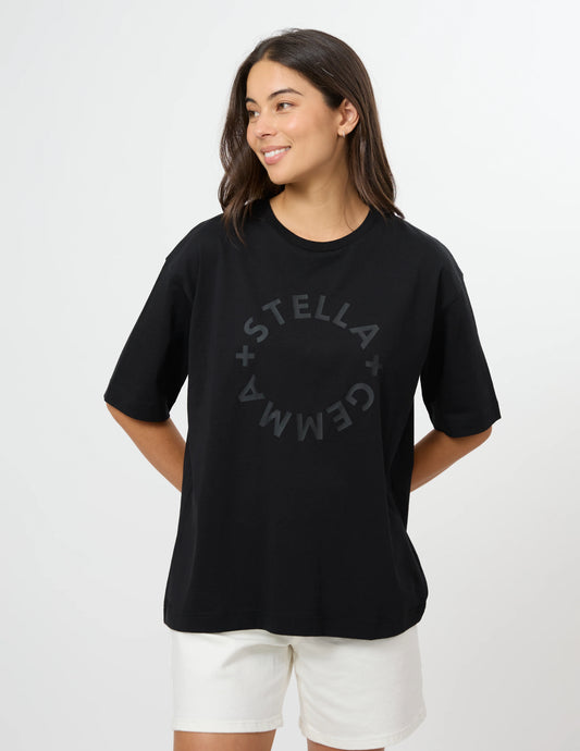 A woman modeling an oversized black t-shirt with a dark gray circular 'STELLA GEMMA' graphic on the chest, paired with white shorts, against a plain white studio background.