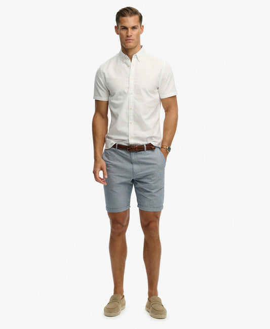 A man modeling a white short-sleeve button-down shirt, light blue chino shorts, a brown leather belt, and beige slip-on loafers against a plain white background.