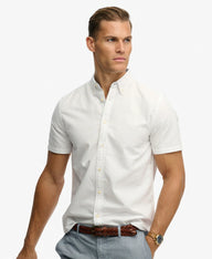 A man wearing a white short-sleeve button-down shirt, styled with light blue jeans, a brown braided belt, and a gold watch, set against a plain white background.
