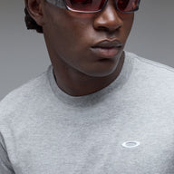 Close-up of a person wearing a light gray heather crewneck t-shirt with a small white Oakley logo on the chest, paired with dark sunglasses, against a neutral gray background.