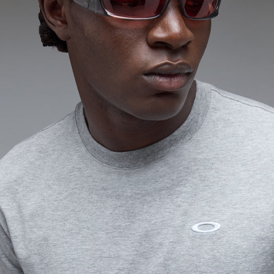 Close-up of a person wearing a light gray heather crewneck t-shirt with a small white Oakley logo on the chest, paired with dark sunglasses, against a neutral gray background.