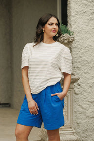 Woman wearing a white ruffled top and blue shorts standing against a stone wall.