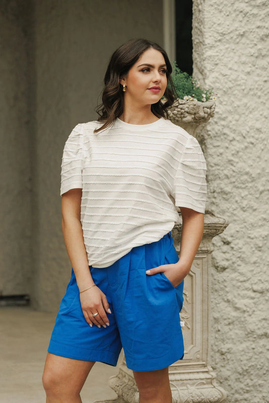 Woman wearing a white ruffled top and blue shorts standing against a stone wall.