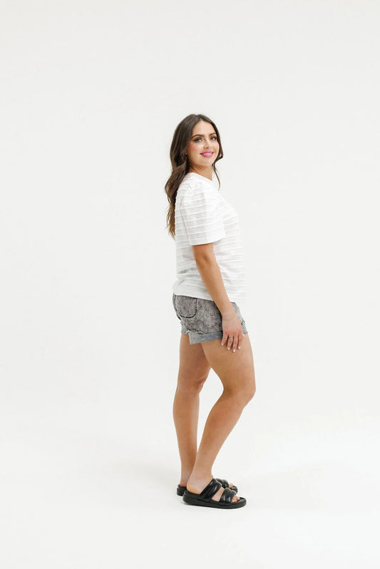A woman modeling a white short-sleeve striped t-shirt, gray denim shorts, and black slide sandals, standing against a solid white studio background.