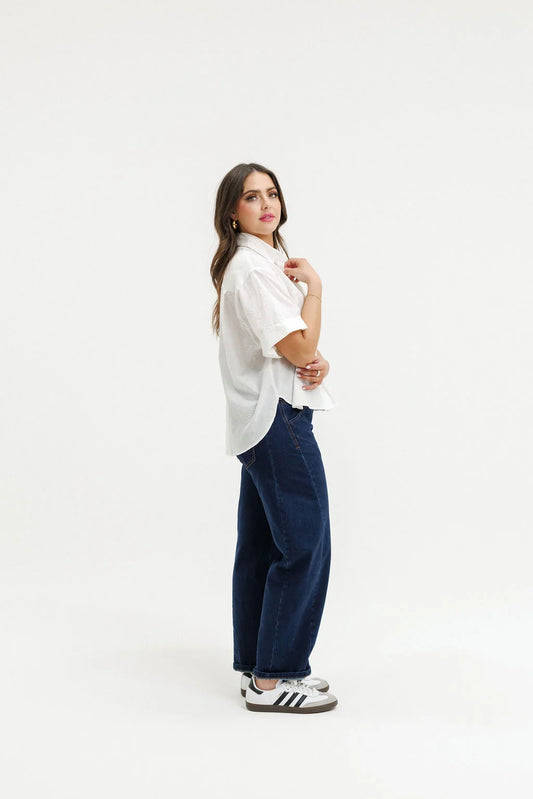 A woman modeling a white short-sleeve button-up shirt and dark blue wide-leg jeans, paired with white sneakers, against a solid white studio background.