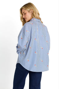 A woman wearing a light blue and white vertically striped button-up shirt with small red and yellow embroidered details, paired with dark blue jeans, shown from a three-quarters back view against a white background.