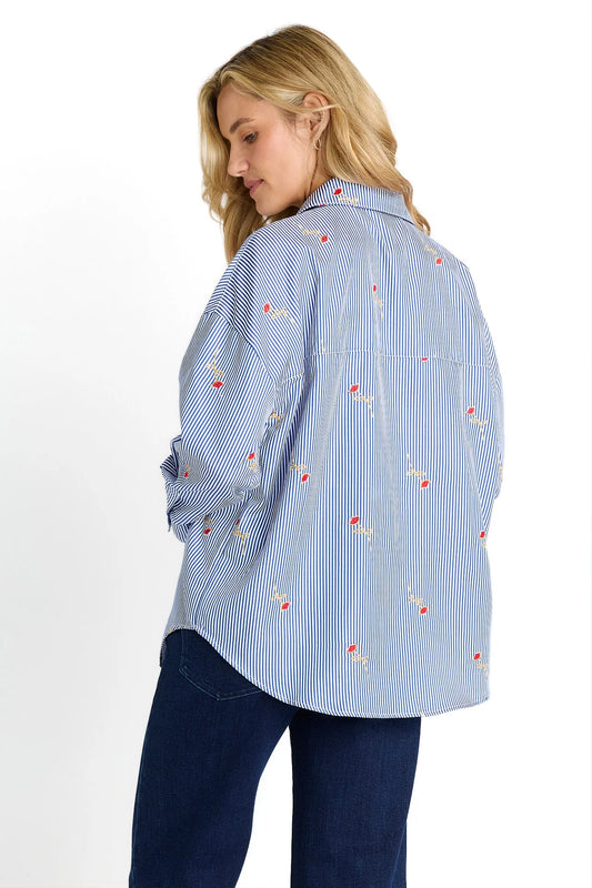 A woman wearing a light blue and white vertically striped button-up shirt with small red and yellow embroidered details, paired with dark blue jeans, shown from a three-quarters back view against a white background.