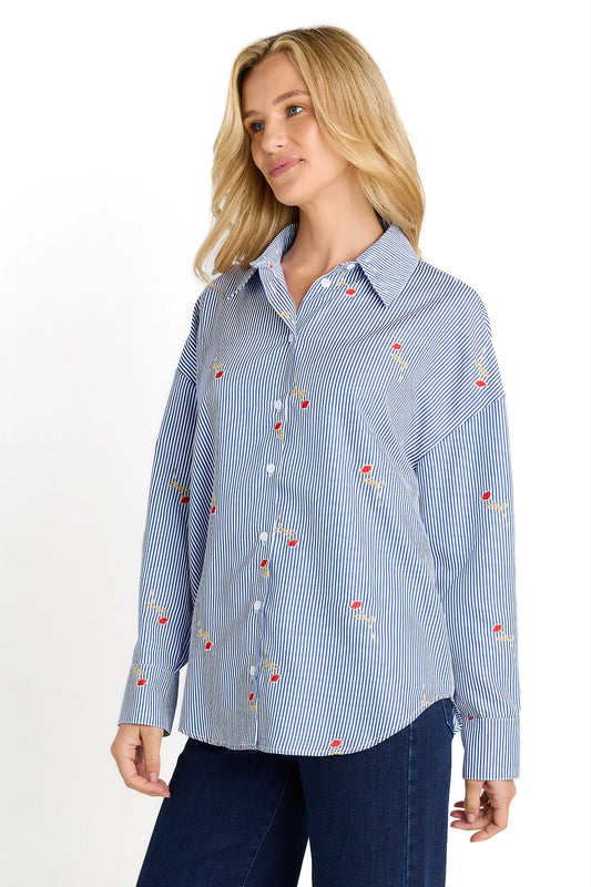 A woman wearing a light blue and white vertically striped button-up shirt with small red floral embroidery, paired with dark blue jeans, against a plain white background.
