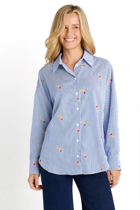 A smiling blonde woman wearing a light blue and white vertically striped button-down shirt with small red and yellow embroidered details, paired with dark blue pants, against a plain white background.