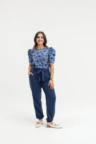 A woman modeling a blue floral puff-sleeve top paired with navy blue trousers and white flat sandals, standing against a plain white studio background.