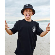 Child wearing a black t-shirt and hat with a logo, giving a thumbs-up on a beach.