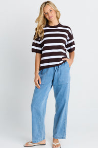 A woman modeling a dark brown and white horizontally striped short-sleeve t-shirt with light blue wide-leg jeans and beige sandals, standing against a light gray studio background.