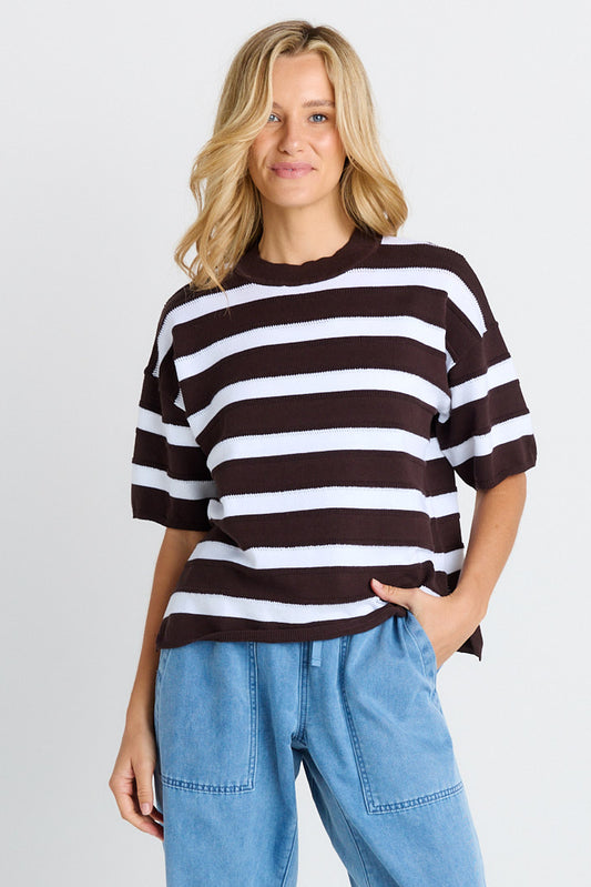 A woman wearing a dark brown and white horizontally striped short-sleeve knit top with light blue jeans, standing against a light gray studio background.