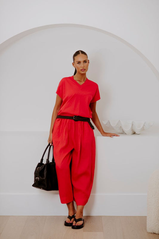A woman modeling a bright red two-piece set consisting of a short-sleeve V-neck top and matching wide-leg cropped pants, styled with a black belt, black sandals, and a black handbag in a minimalist white room.