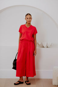 A woman modeling a bright red two-piece linen set consisting of a short-sleeve V-neck top and matching wide-leg cropped pants, styled with black sandals and a black handbag in a minimalist white room with an arched wall.