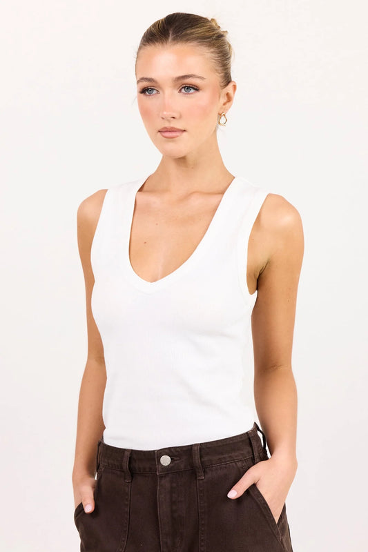 A woman modeling a white ribbed sleeveless tank top with a deep V-neck, paired with dark brown pants, against a plain white studio background.