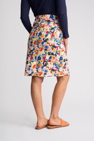 A model is shown from the back wearing a colorful, high-waisted A-line skirt with a dense abstract floral pattern, paired with a navy blue ribbed top and peach-colored slide sandals, against a light gray studio background.