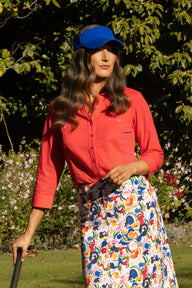 A woman outdoors wearing a bright coral-red long-sleeve button-up shirt, a blue visor, and a white midi skirt with a colorful abstract print. She is standing in a grassy garden with trees in the background.