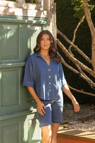 A woman wearing a blue denim short-sleeve button-up shirt and matching denim shorts, standing outdoors next to a green wooden door with branches and greenery in the background.
