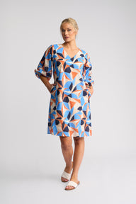 A woman modeling a short-sleeve V-neck dress with a colorful geometric triangle pattern in blue, orange, navy, and beige, paired with white slide sandals, against a light gray studio background.