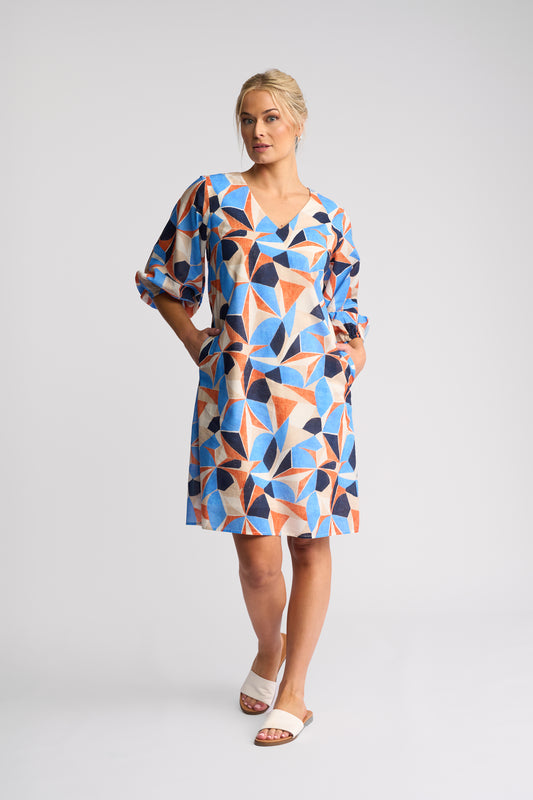 A woman modeling a short-sleeve V-neck dress with a colorful geometric triangle pattern in blue, orange, navy, and beige, paired with white slide sandals, against a light gray studio background.