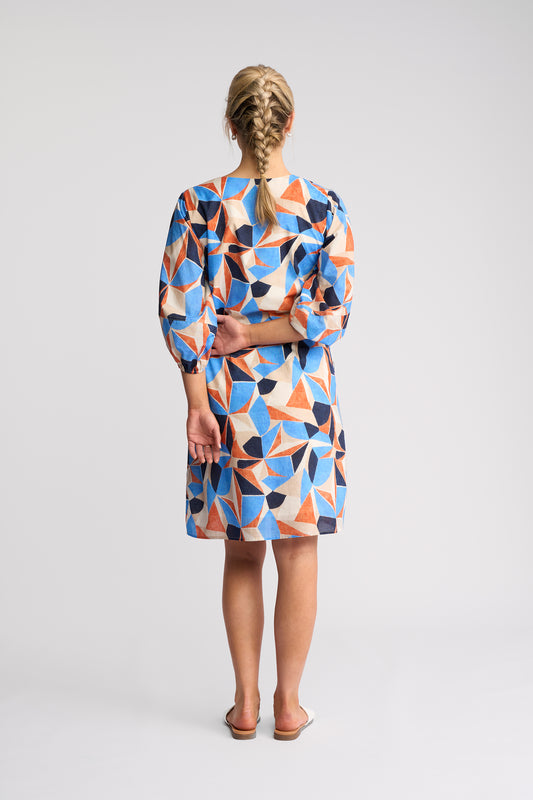 A woman viewed from behind wearing a knee-length dress with a colorful geometric pattern in blue, orange, beige, and black, paired with brown sandals, against a light gray studio background.