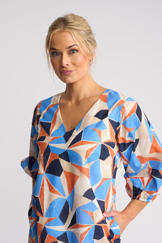 A woman wearing a loose-fitting V-neck blouse with a bold geometric starburst pattern in blue, orange, navy, and cream, photographed against a light gray studio background.