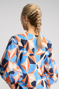A woman shown from the back wearing a loose-fitting blouse with a bold geometric starburst pattern in blue, orange, beige, and navy on a light gray studio background.