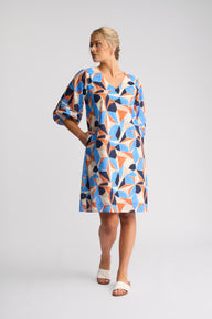 A woman modeling a short-sleeve V-neck dress with a colorful geometric triangle pattern in blue, orange, navy, and cream, paired with white slide sandals, against a light gray studio background.