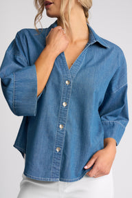 A woman wearing a blue denim button-up shirt with three-quarter sleeves and white pants, shown from the neck down against a light gray studio background.
