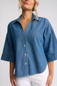 A woman wearing a blue denim button-up shirt with three-quarter sleeves and white pants, shown from the neck down against a light gray studio background.